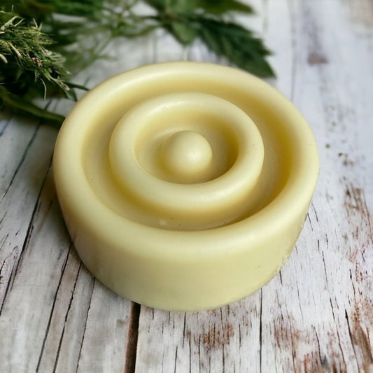 Cream coloured solid lotion bar with concentric ring design on whitewashed background with herbs in the background
