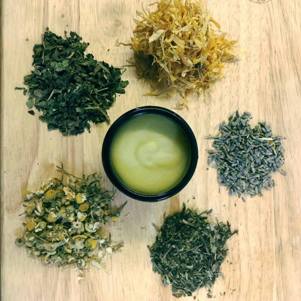 Cream coloured soothing botanical balm in amber jar with dried herbs of lavender, comfrey, chamomile, plantain and calendula around on chopping board