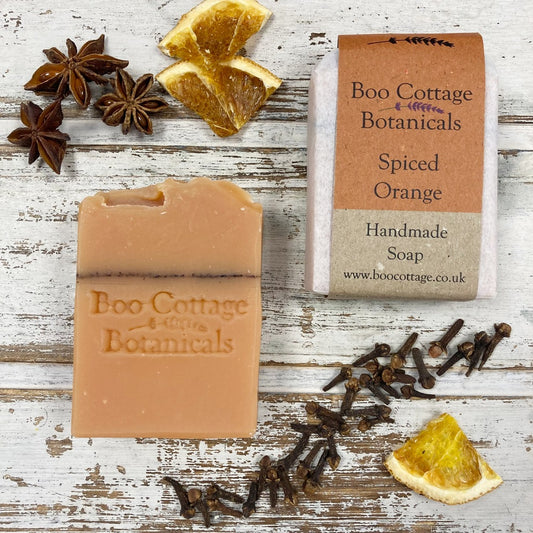 Orange soaps, one unwrapped, one with cardboard label on whitewashed background with dried oranges, cloves and star anise scattered around