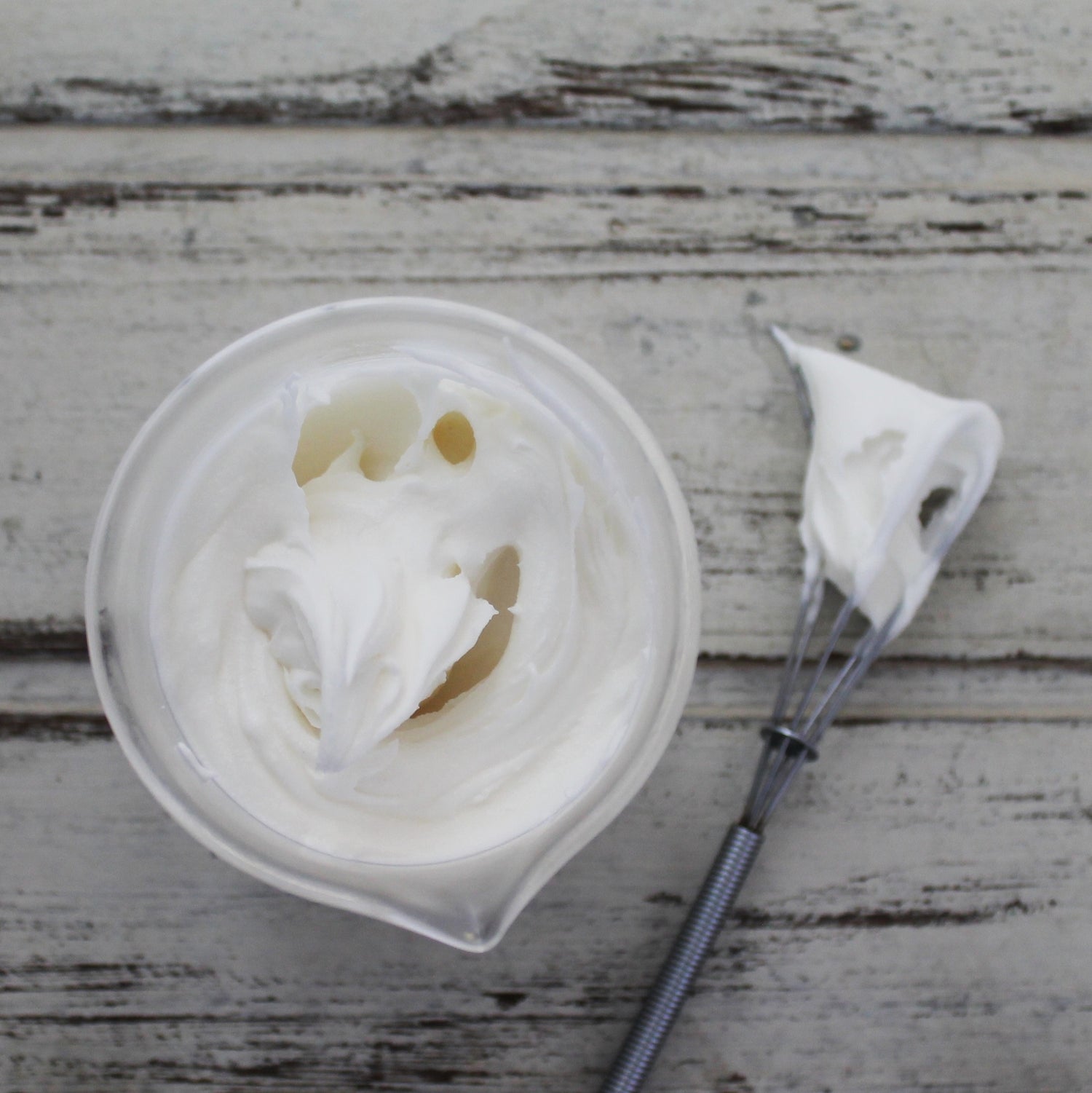 White whipped body butter in glass beaker with mini whisk on whitewashed wooden background