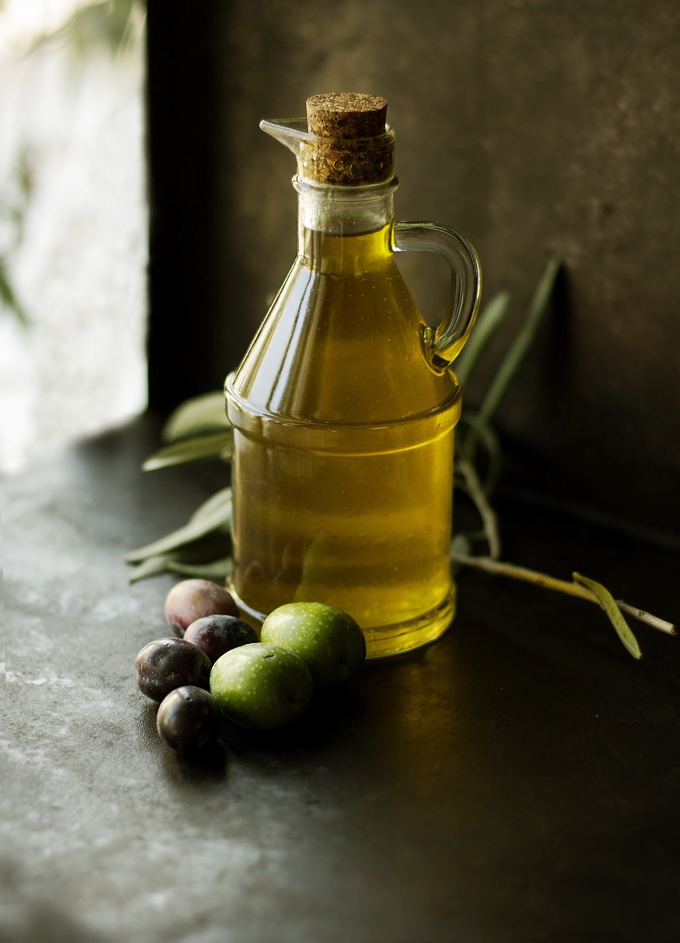 Golden olive oil in bottle with fresh olives on dark table