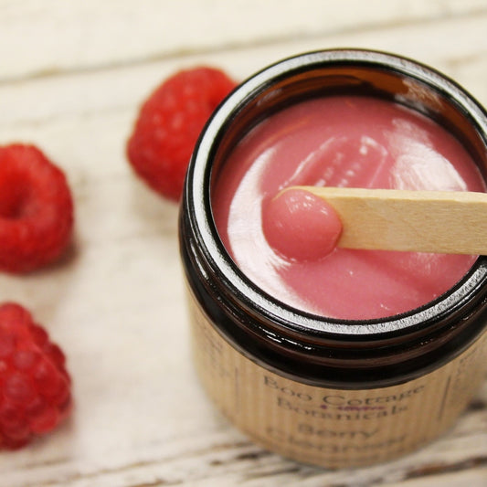 Glossy pink balm in amber jar being scooped with wooden spatula on white background with raspberries