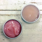 Mini aluminium tins of berry cleanser showing difference in colour between bright and pale pink on white washed wooden background