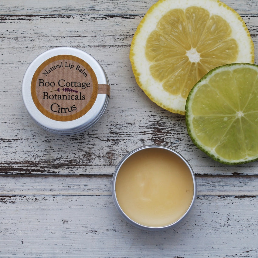 Yellow lip balm in small tin, one open, one closed with lemon and lime slice on whitewashed wooden background 