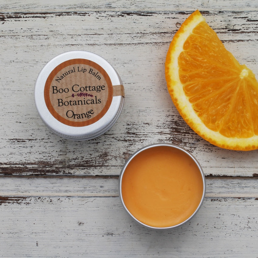 Orange lip balm in open tin and lidded tin on whitewashed wooden background with orange slice