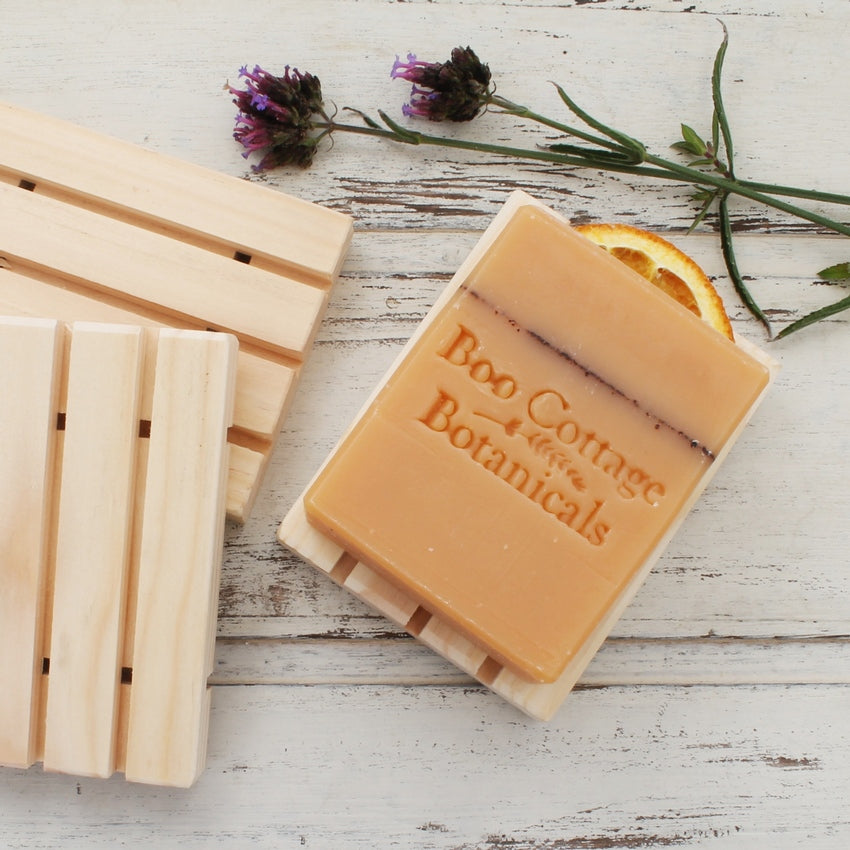 Orange soap bar on pale wooden soap dish with other soap dishes and flowers on whitewashed wooden background