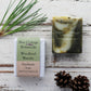 Dark green and cream marbled soap bar with wrapped bar next to it, with pine needles and pine cones