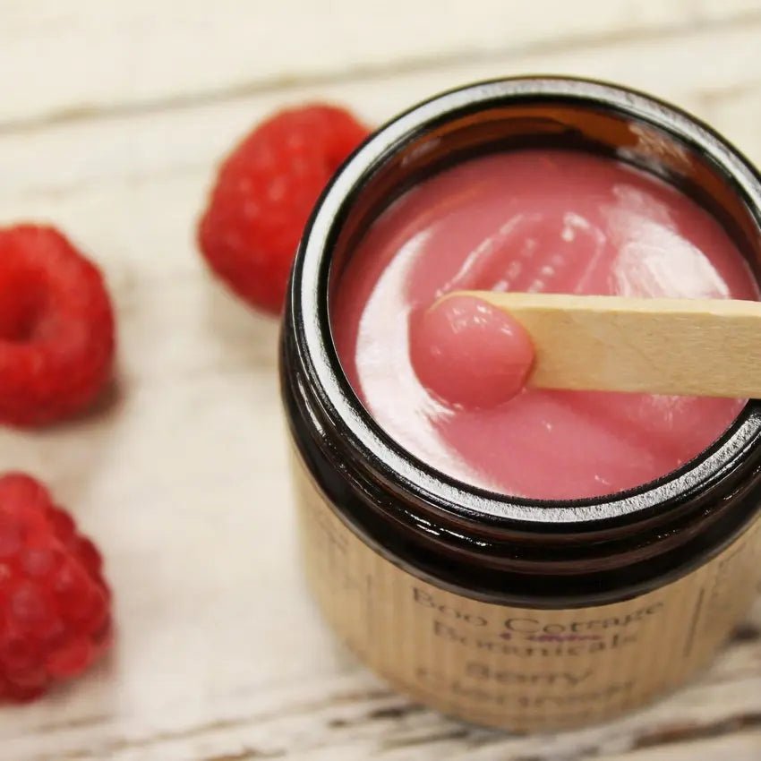 Amber glass jar with glossy pink balm being scooped with wooden spatula with raspberries on white background