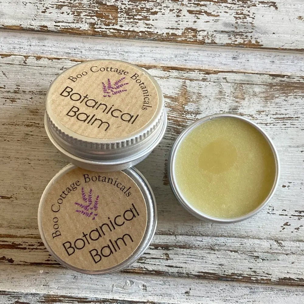 Pile of 3 mini tins of botanical balm, one with open lid showing cream colour of balm on whitewashed wooden background