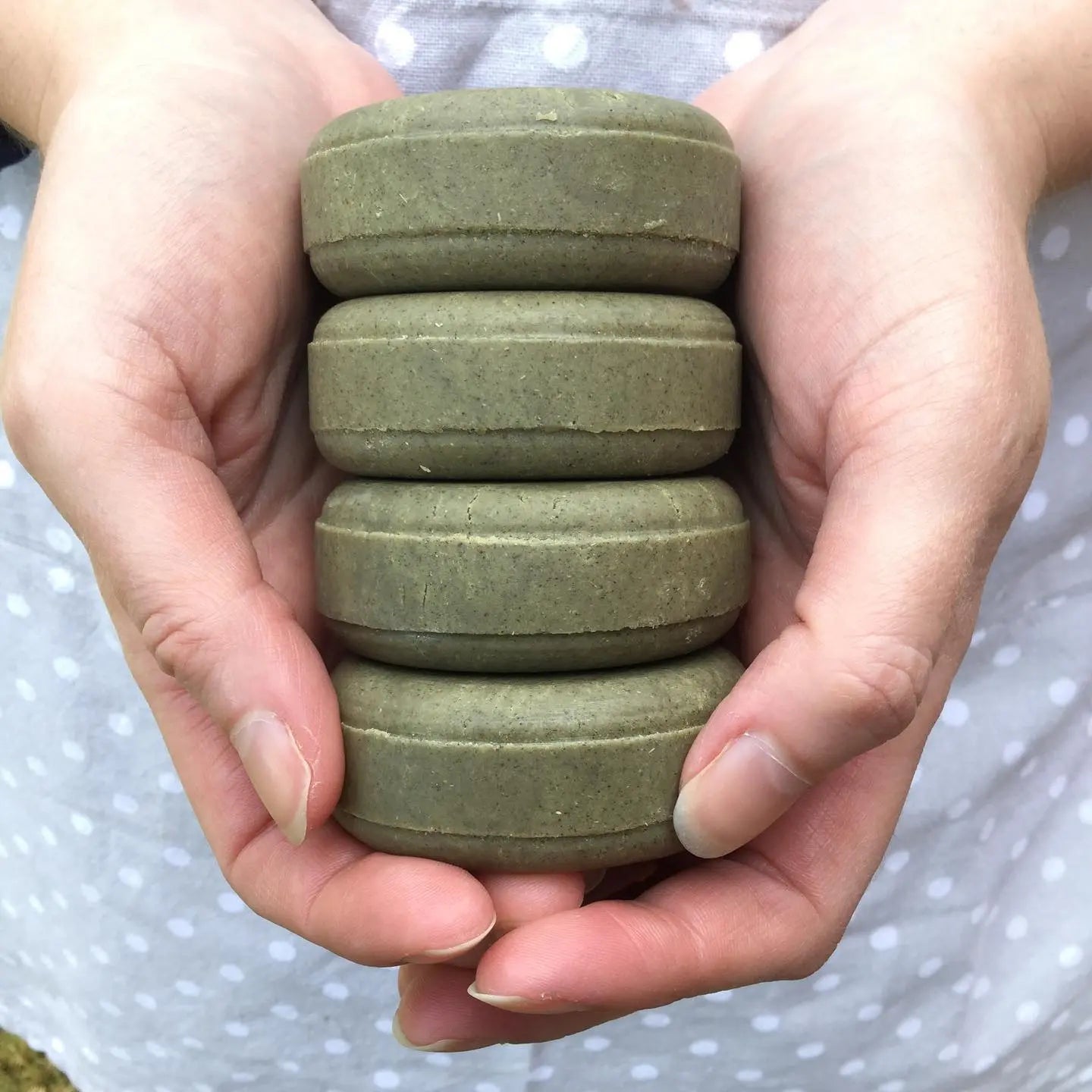 Stack of 4 green rround shampoo bars in white hands with grey spotty apron background