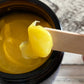 Close up view of yellow balm on wooden spatula with open amber jar on white wooden background