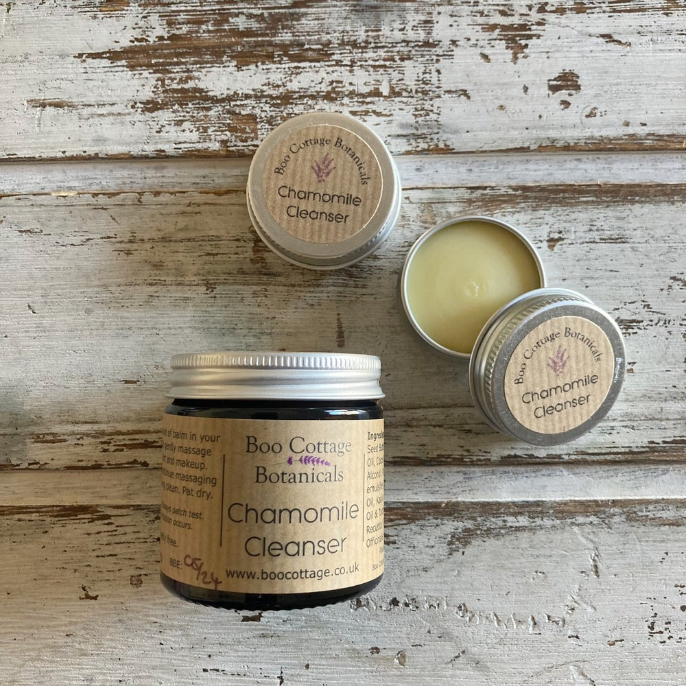 Amber jar with brown label and aluminium lid of chamomile cleanser alongside mini aluminium tins on white background