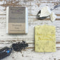 Cream speckled soap bar with grey card wrapped bar, poppy seeds and cream mango butter on spoon on white wood background