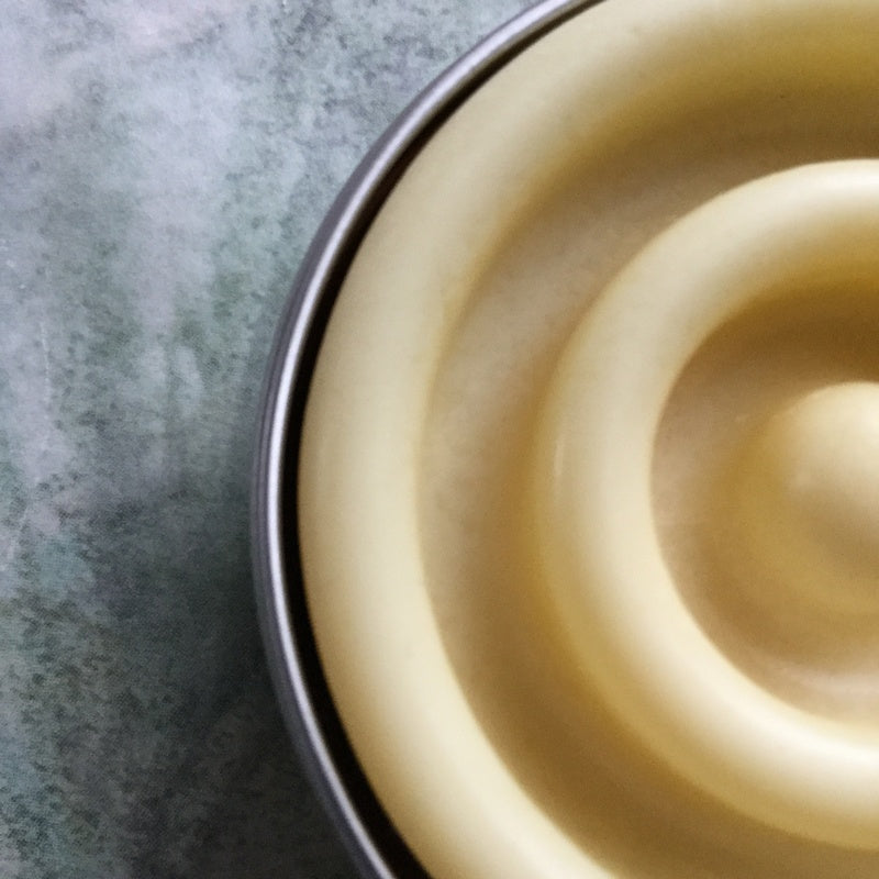 Close up view of curved texture on surface of solid lotion bar in tin with mottled green background