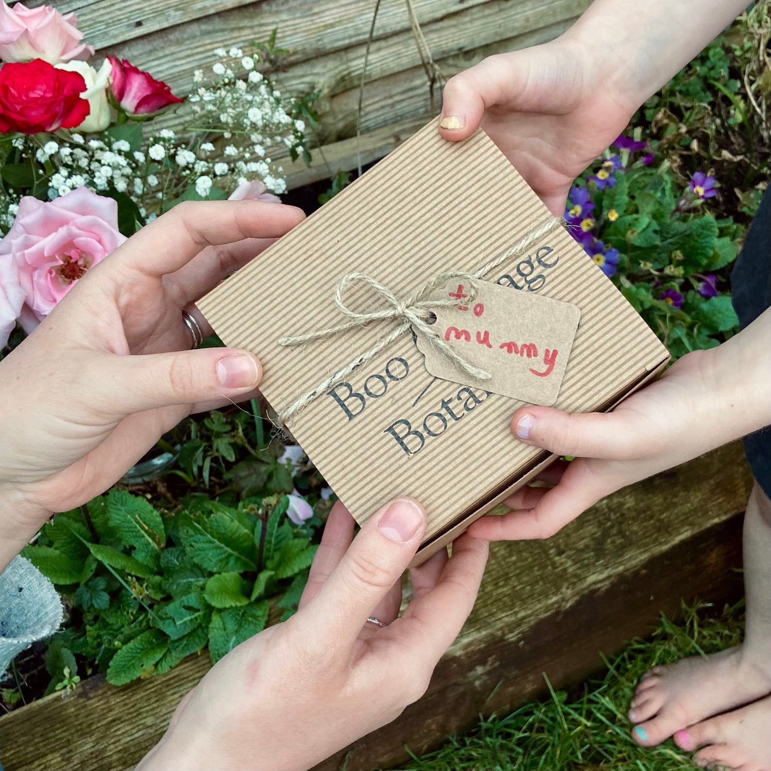 A brown gift box with a tag saying 'to mummy' being handed over between a child with colourful finger nails and a mum's hands in a garden with flowers behind.