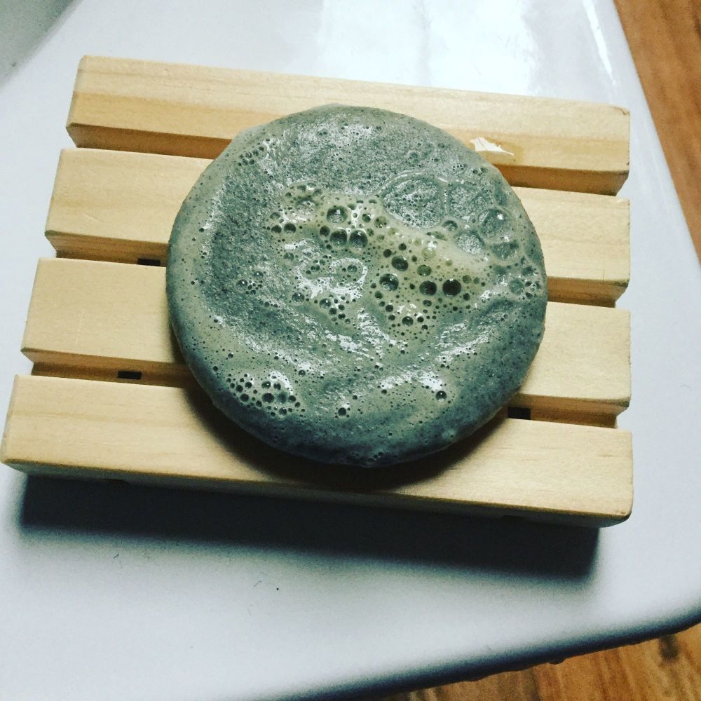 Bubbly natural green round shampoo bar on wooden soap dish sat on side of white bath tub
