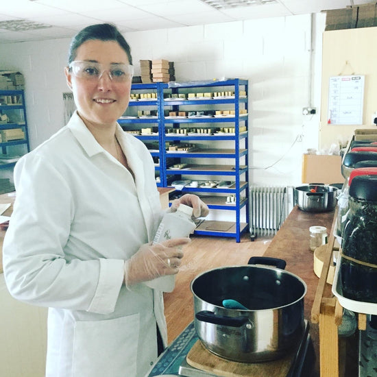 Smiling brunette female in white lab coat and safety goggles with saucepan in front and shelves full of soap behind