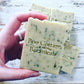 Pale green bar of soap with dark green flecks held in hand with more bars behind on white wood background