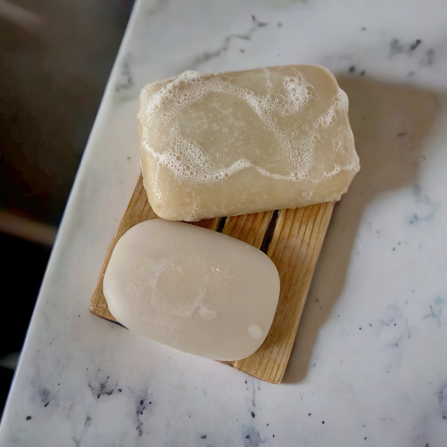 Two natural coloured soaps on a natural wooden soap dish on marble counter