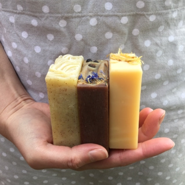Cream speckled, dark brown and yellow soaps with botanical toppings in white female hand with grey spotty apron behind
