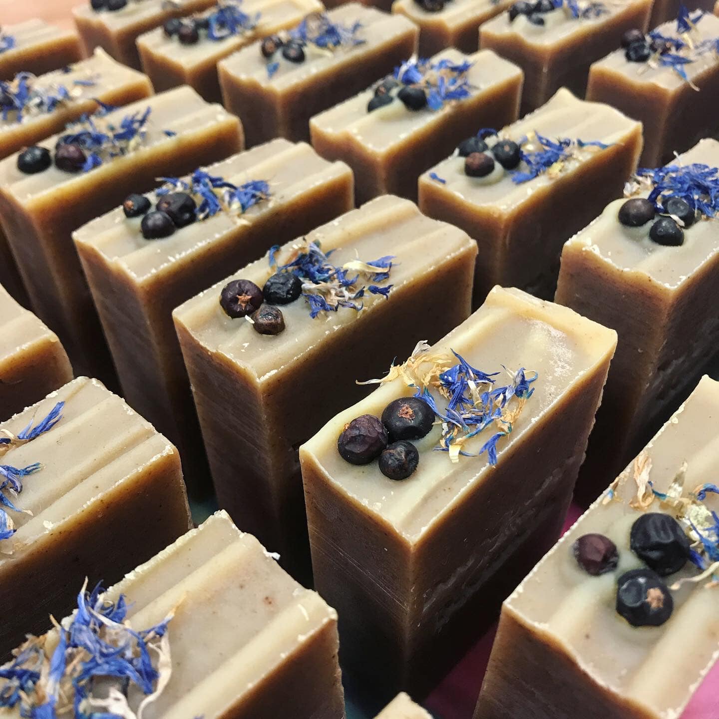 Collection of brown soaps with blue cornflowers and black juniper berries on top or each bar spread out to dry