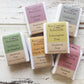 Six bars of different natural soap wrapped in multicoloured card on white wood background