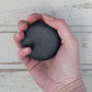 Black flat round shampoo bar in white female hand with whitewashed wooden background