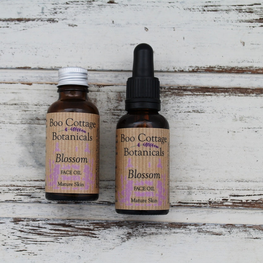 Amber bottles of Blossom face oil, one with black pipette lid, one with aluminium lid on whitewashed wooden background