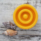 Close up view of round yellow solid lotion bar showing textured rings on whitewashed wooden background with sea shells