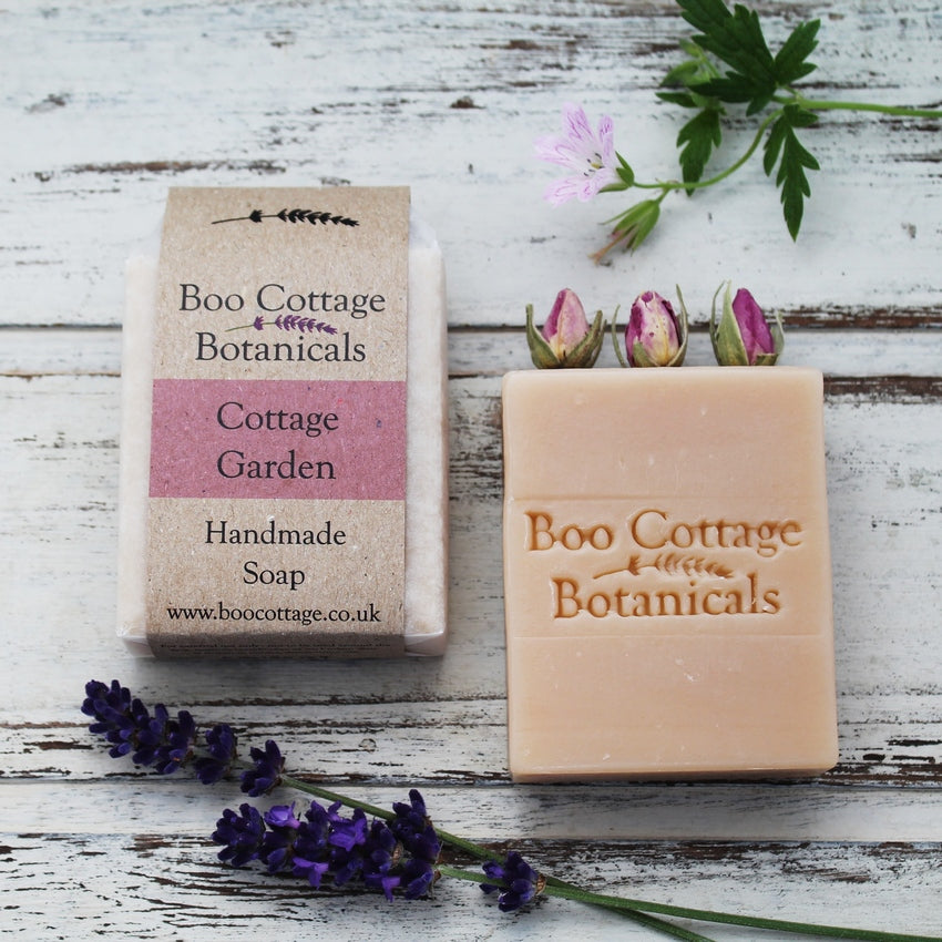 Wrapped and unwrapped pink soap bar with rose buds on top on whitewashed wooden background with lavender and geranium flowers