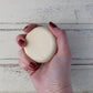Cream flat round shampoo bar in white female hand with dark red nails on whitewashed wooden background