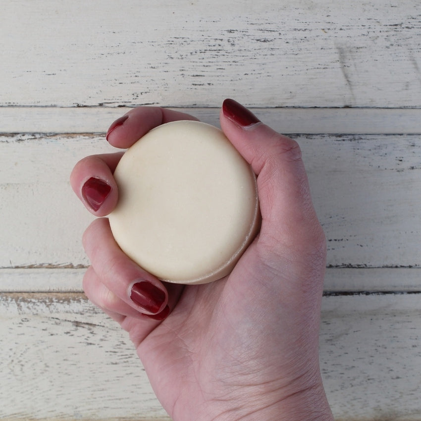 Cream flat round shampoo bar in white female hand with dark red nails on whitewashed wooden background