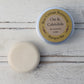 Cream flat round shampoo bar unwrapped and in aluminium tin on whitewashed wooden background