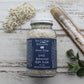 Bottle of pale green bath salts with navy blue label on whitewashed wooden background with salt, mint leaves and sea shell