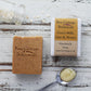 Unwrapped and wrapped brown speckled soap on whitewashed wooden background with spoon of honey, jar of oats