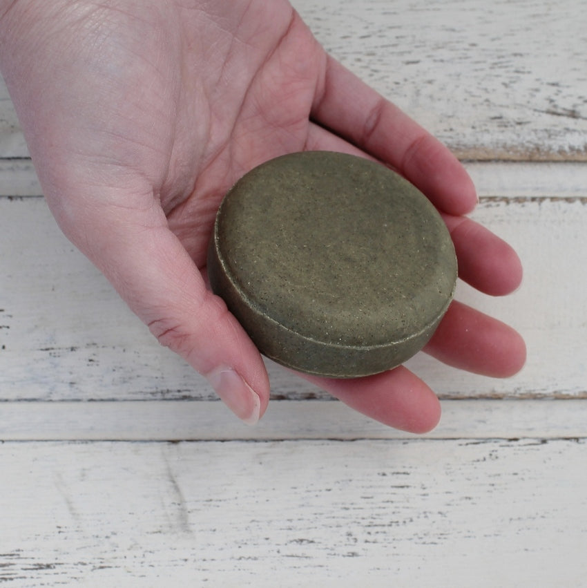 Dark green flat round shampoo bar in female white hand with whitewashed wooden background