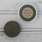 Dark green round shampoo bar unwrapped and in aluminium tin on whitewashed wooden background