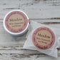 Meadow lotion bar in aluminium tin and in waxed paper bag with pink labels on whitewashed wooden background