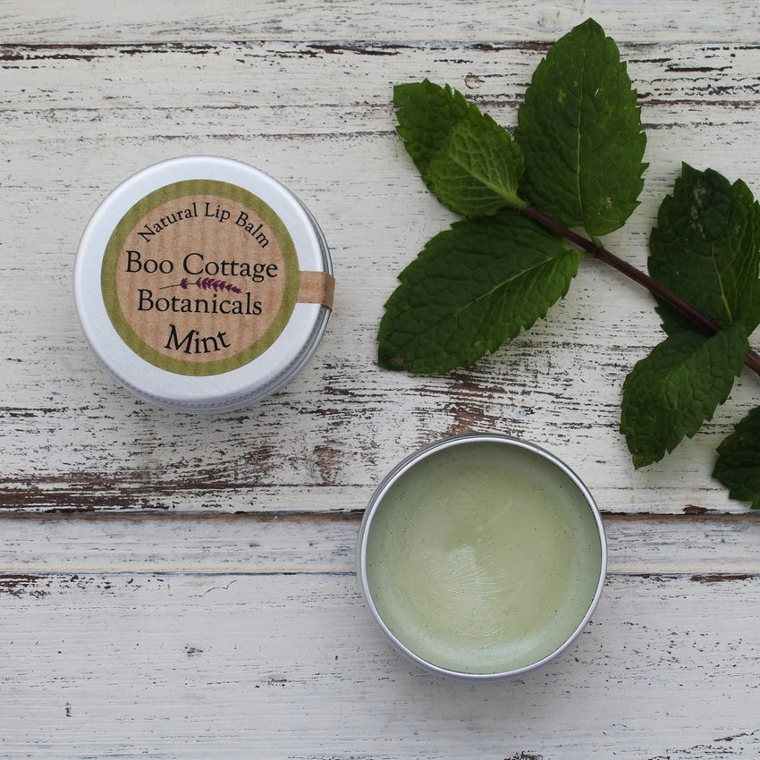 Green lip balm with open tin and lidded round tin on whitewashed wooden background with mint leaves