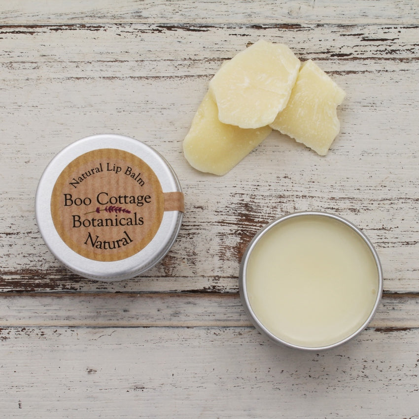 Cream lip balm with open tin and lidded round tin on whitewashed wooden background with cream pieces of cocoa butter