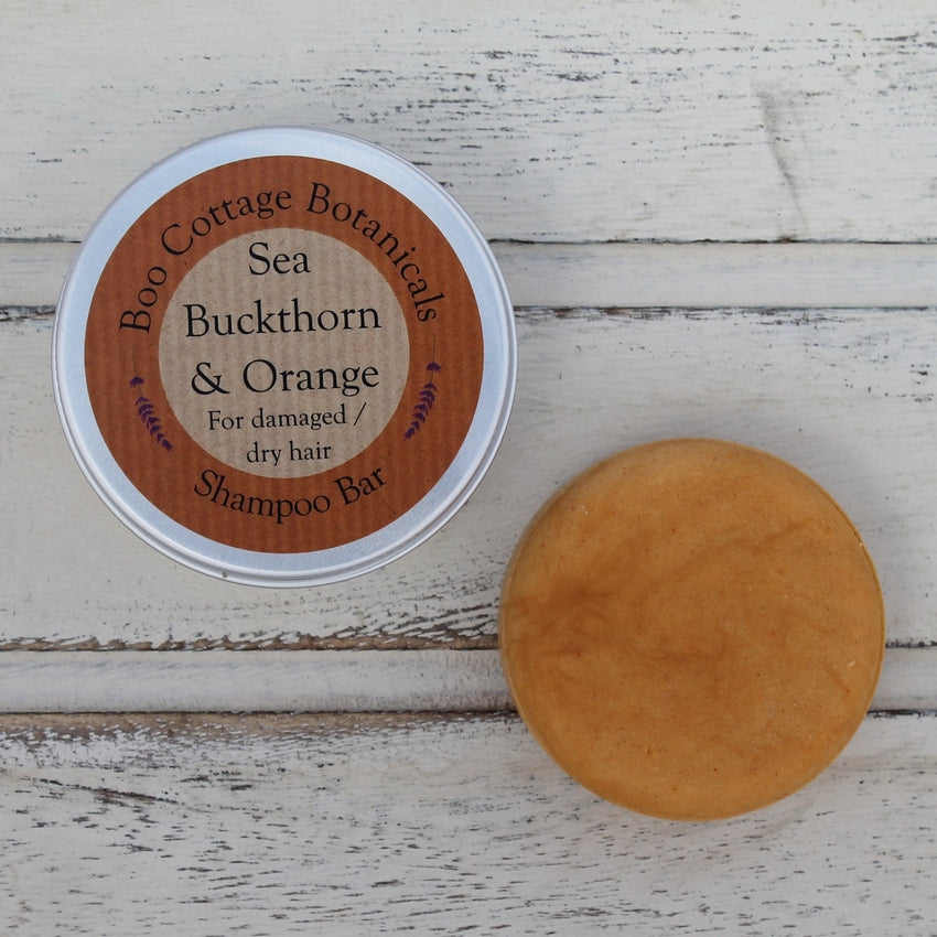 Orange flat round shampoo bar unwrapped and aluminium tin on whitewashed wooden background