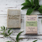 Unwrapped and wrapped green speckled soap bars with mint and nettle leaves on whitewashed wooden background