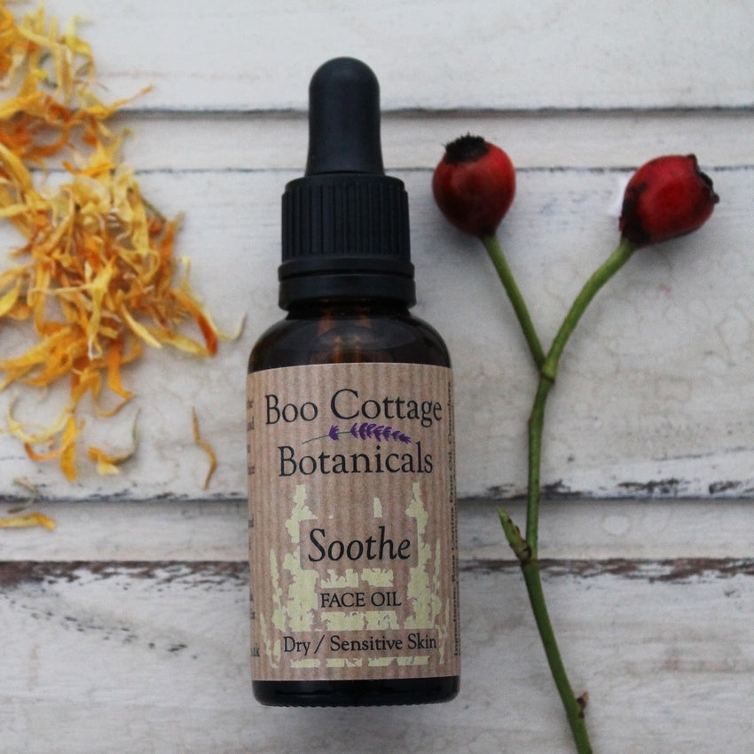 Amber bottle of Soothe face oil with black dropper pipette with rosehips and dried calendula petals on whitewashed wooden background