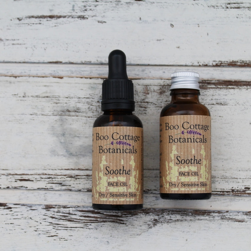 Amber bottles of Soothe face oil, one with black dropper pipette and one with aluminium lid on whitewashed wooden background