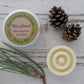 Round aluminium tin with green label with round, cream, textured solid lotion bar on whitewashed background with pine needs and pine cones