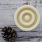 Cream, round textured solid lotion bar on whitewashed wooden background with pine cone